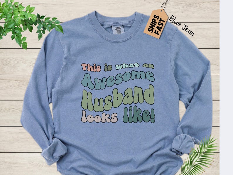May include: A light blue long-sleeved t-shirt with the text "This is what an Awesome Husband looks like!" in colorful, retro-style lettering.