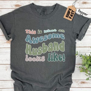 May include: A dark gray t-shirt with the text "This is what an Awesome Husband looks like!" in colorful, retro-style lettering.