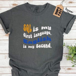 May include: Dark gray t-shirt with rolled sleeves. The shirt features the text "SQL is my first language, English is my second" in orange, white, and blue. A small tag with "SHIPS FAST" is attached to the shirt.