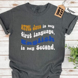 May include: A dark gray t-shirt with the text "HTML Java is my first language, English is my second." printed in white, blue, and orange.