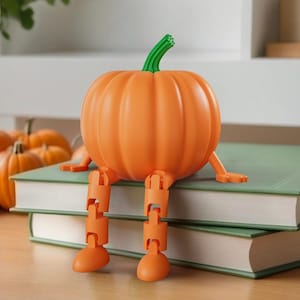 Puede incluir: Un sujetalibros con forma de calabaza naranja con patas articuladas y un tallo verde se asienta sobre dos libros verdes. La calabaza es de color naranja brillante, perfecta para la decoración de otoño. Otras calabazas son visibles en el fondo.
