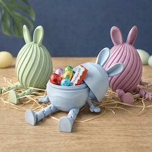 May include: Three Easter bunny-shaped containers in pastel colors: light green, light blue, and pink. The light blue bunny is open, revealing colorful foil-wrapped chocolate eggs and a candy bar. The bunnies sit on a bed of straw, with a blue background.