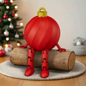 以下が含まれることがあります： 木製の丸太に座っている、関節式の脚と腕を持つ、気まぐれな赤いクリスマスオーナメントのフィギュア。オーナメントは金色のキャップが付いており、丸いニュートラルカラーのラグの上に置かれています。背景にはクリスマスツリーが見えます。