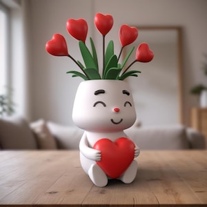Könnte beinhalten: Ein weißer Keramik-Pflanztopf in Form eines lächelnden Gesichts, der ein rotes Herz in seinen Händen hält. Der Topf hat schwarze Augen und eine rote Nase. Der Topf ist mit grünen Blättern und roten herzförmigen Blumen gefüllt.