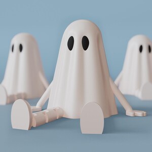 Op de afbeelding: Drie witte spookfiguren met zwarte ovale ogen en uitgestrekte armen zitten tegen een lichtblauwe achtergrond. De geesten hebben een gedrapeerd ontwerp en zitten met hun benen uitgestrekt. De figuren zijn een grillig, minimalistisch ontwerp.