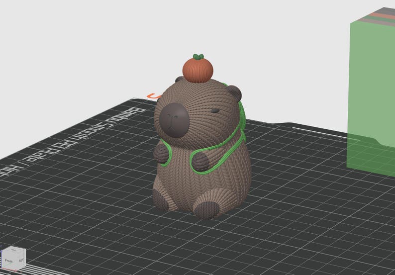 May include: A brown and green 3D printed capybara figurine wearing a green scarf and holding an orange pumpkin on its head.