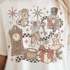 May include: A cream-colored t-shirt featuring a graphic print of Nutcracker-themed illustrations, including a mouse king, a nutcracker soldier, a ballerina, ballet slippers, a grandfather clock, a drum, a gift box, and a key, accented by stars.