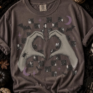 Op de afbeelding: Een antracietgrijs T-shirt met korte mouwen en een afbeelding van twee bleke handen die een hartvorm maken. Het ontwerp bevat een spin, vleermuizen, sterren, maansikkels en een verweerde zwart-witte schaakbordpatroon achtergrond.