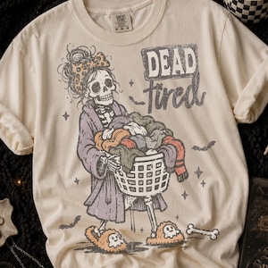 May include: A cream-coloured t-shirt featuring a graphic of a skeleton wearing a purple robe, a leopard print hair bow, and ghost-shaped slippers. The skeleton holds a laundry basket filled with clothes. The text reads DEAD tired.