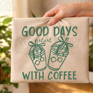 K&ouml;nnte beinhalten: Beige Tragetasche mit gr&uuml;nem Text "GOOD DAYS start WITH COFFEE". Das Design umfasst zwei gr&uuml;ne Kaffeetassen-Illustrationen mit Eisw&uuml;rfeln und Schleifen. Die Tasche wird von einer Hand gehalten.