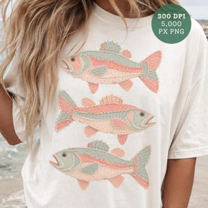 Puede incluir: Camiseta de color crema con tres diseños de peces estilizados. Los peces tienen acentos rosas, grises y melocotón, con costuras detalladas. La camiseta tiene un corte holgado y un cuello redondo. La imagen también incluye un círculo verde con el texto "300 DPI 5,000 PX PNG".