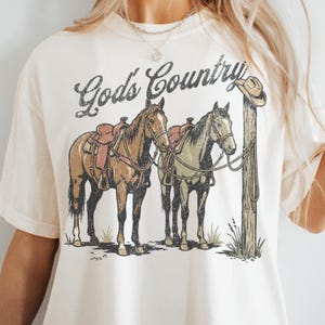 May include: A cream-colored t-shirt featuring a graphic print of two saddled horses tied to a wooden post with a cowboy hat hanging on top. The text God's Country is printed in a dark, distressed script font above the horses in the center.