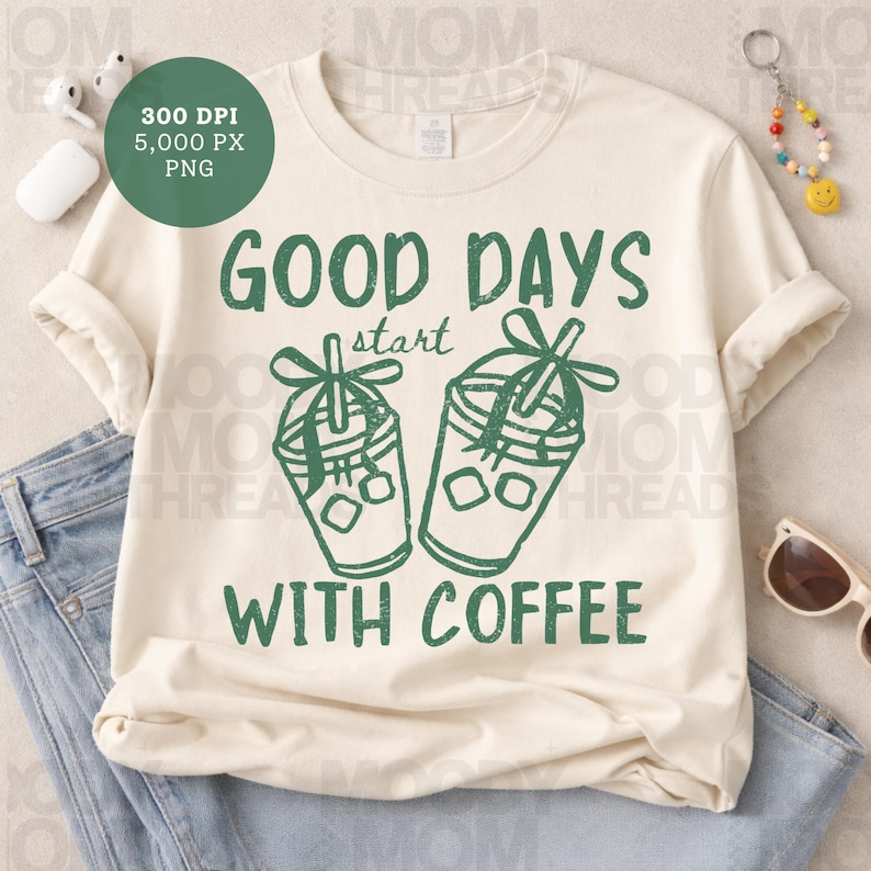 K&ouml;nnte beinhalten: Cremefarbenes T-Shirt mit dem gr&uuml;nen Text "GOOD DAYS start WITH COFFEE". Das Design umfasst zwei gr&uuml;ne Kaffeetassen-Illustrationen mit Eisw&uuml;rfeln und Strohhalmen. Das Shirt ist gefaltet und zeigt die &Auml;rmel und einen Teil der Jeans.