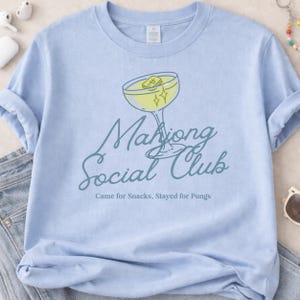 K&ouml;nnte beinhalten: Hellblaues T-Shirt mit einer Cocktail-Grafik und dem Text "Mahjong Social Club". Der Cocktail ist gelb mit einem Mahjong-Stein und Sternen. Der Text "Came for Snacks, Stayed for Pungs" steht darunter.