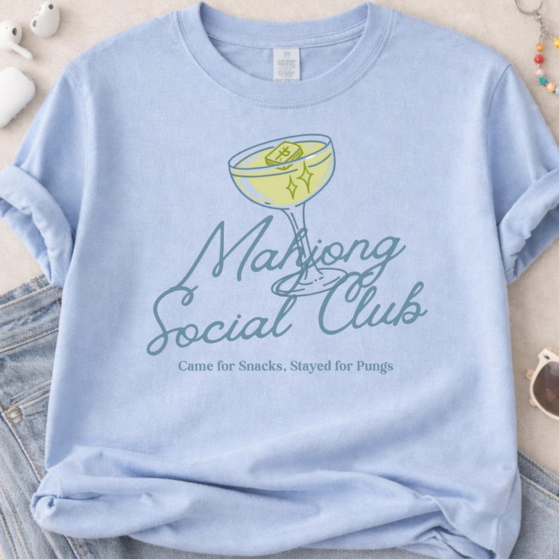 K&ouml;nnte beinhalten: Hellblaues T-Shirt mit einer Cocktail-Grafik und dem Text "Mahjong Social Club". Der Cocktail ist gelb mit einem Mahjong-Stein und Sternen. Der Text "Came for Snacks, Stayed for Pungs" steht darunter.