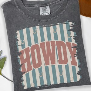 Peut inclure: T-shirt gris Comfort Colors avec un motif vieilli &laquo; HOWDY &raquo; en lettres rouges. Le motif pr&eacute;sente des rayures bleues et blanches avec des fers &agrave; cheval. Le t-shirt est pli&eacute;.