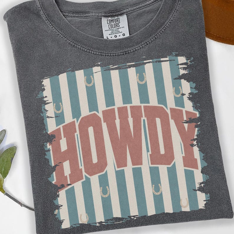 Peut inclure: T-shirt gris Comfort Colors avec un motif vieilli &laquo; HOWDY &raquo; en lettres rouges. Le motif pr&eacute;sente des rayures bleues et blanches avec des fers &agrave; cheval. Le t-shirt est pli&eacute;.