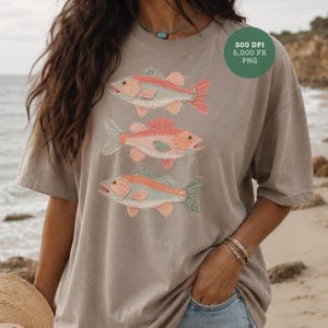 Puede incluir: Camiseta beige con una ilustración detallada de tres peces en tonos rojos, melocotón y azul verdoso. El texto "300 DPI 5,000 PX PNG" está en un círculo verde en la parte superior derecha.