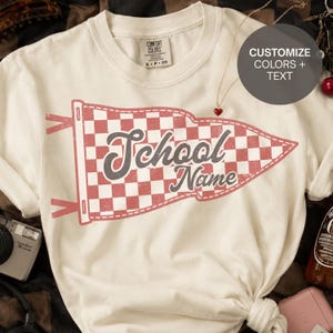 Può includere: Una t-shirt a maniche corte color crema con una grande grafica di un gagliardetto a scacchi rossi e bianchi dall'aspetto vissuto. Il testo "School Name" è stampato in un carattere corsivo grigio scuro al centro del gagliardetto. Un sovrapposizione circolare riporta "CUSTOMIZE COLORS + TEXT".