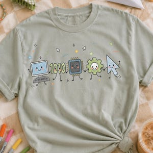 May include: A sage green crew neck t-shirt featuring a graphic of a computer, the numbers 1010, a microchip, a gear, and a cursor holding hands. Each element has a smiling face, and the design includes small stars and cursor icons.