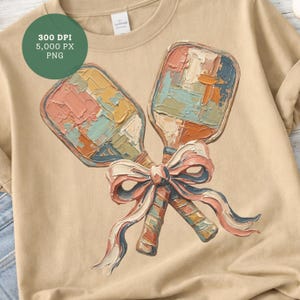 May include: A beige t-shirt featuring a colorful illustration of two pickleball paddles tied together with a ribbon. The paddles have a textured, painted appearance with shades of pink, blue, orange, and yellow. The ribbon is white, pink, and blue.
