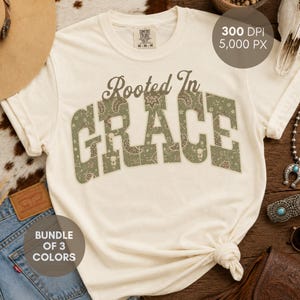 May include: A cream-colored short-sleeve t-shirt featuring the text Rooted In GRACE in a large, block-letter font. The letters are filled with a sage green pattern of paisley, stars, and cow skulls. The shirt has a crew neckline and a tag.