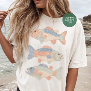 Puede incluir: Camiseta color crema con tres diseños de peces coloridos. Cada pez es un mosaico de telas azules, melocotón y verde claro. La camiseta también tiene un círculo verde con el texto "300 DPI 5,000 PX PNG".