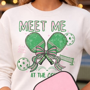 May include: White sweatshirt with a green and pink graphic. The graphic says "MEET ME" above two green pickleball paddles tied with a pink bow. The words "dink, drop, drive" are written on the left side. The words "AT THE CO" are at the bottom.