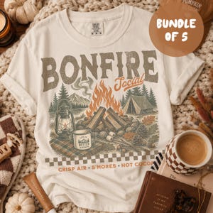 May include: An off-white t-shirt featuring a graphic of a campfire, a tent, a lantern, and a mug with the text 'LIFE IS BETTER'. The shirt displays the text 'BONFIRE SOCIAL' and 'CRISP AIR S'MORES HOT COCOA' with a checkered border at the bottom.