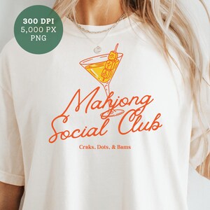 K&ouml;nnte beinhalten: Cremefarbenes T-Shirt mit dem Schriftzug "Mahjong Social Club" in orangefarbener Schrift. Eine Cocktailglas-Grafik mit einem Spie&szlig; mit Mahjong-Steinen befindet sich &uuml;ber dem Text. Darunter steht "Craks, Dots, & Bams".