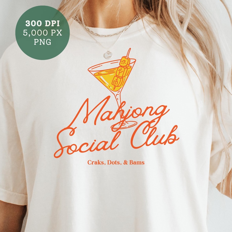K&ouml;nnte beinhalten: Cremefarbenes T-Shirt mit dem Schriftzug "Mahjong Social Club" in orangefarbener Schrift. Eine Cocktailglas-Grafik mit einem Spie&szlig; mit Mahjong-Steinen befindet sich &uuml;ber dem Text. Darunter steht "Craks, Dots, & Bams".
