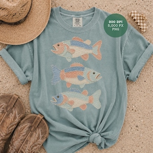 Puede incluir: Una camiseta azul claro con tres diseños de peces. Cada pez tiene un diseño de patchwork con colores azul, melocotón y blanco. La camiseta está combinada con un sombrero de paja y botas de vaquero.