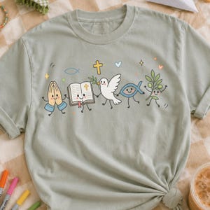 May include: A sage green short-sleeve t-shirt featuring a graphic print of five anthropomorphic characters holding hands: praying hands, an open Bible, a white dove with an olive branch, a blue fish, and a green olive branch with a smiling face.
