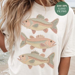 May include: Cream-colored t-shirt featuring three embroidered fish in pastel colors. The fish have pink, green, and yellow details, with open mouths and detailed fins. The shirt has a relaxed fit and a round neckline.