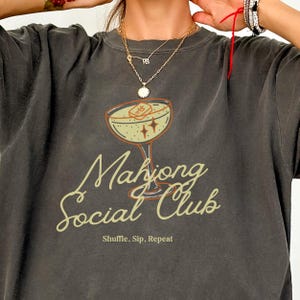 K&ouml;nnte beinhalten: Dunkelgraues T-Shirt mit einem Cocktailglas-Motiv. Das Glas enth&auml;lt einen Mahjong-Stein und Sterne. Die Worte "Mahjong Social Club" sind in einer Schreibschrift geschrieben, darunter "Shuffle, Sip, Repeat".