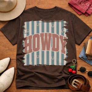 Peut inclure: Un t-shirt marron avec le mot "HOWDY" dans une police de style western vieillie. Le design comprend des rayures bleues et blanches et des motifs de fers &agrave; cheval. Fait partie d'une collection de v&ecirc;tements sur le th&egrave;me du western.