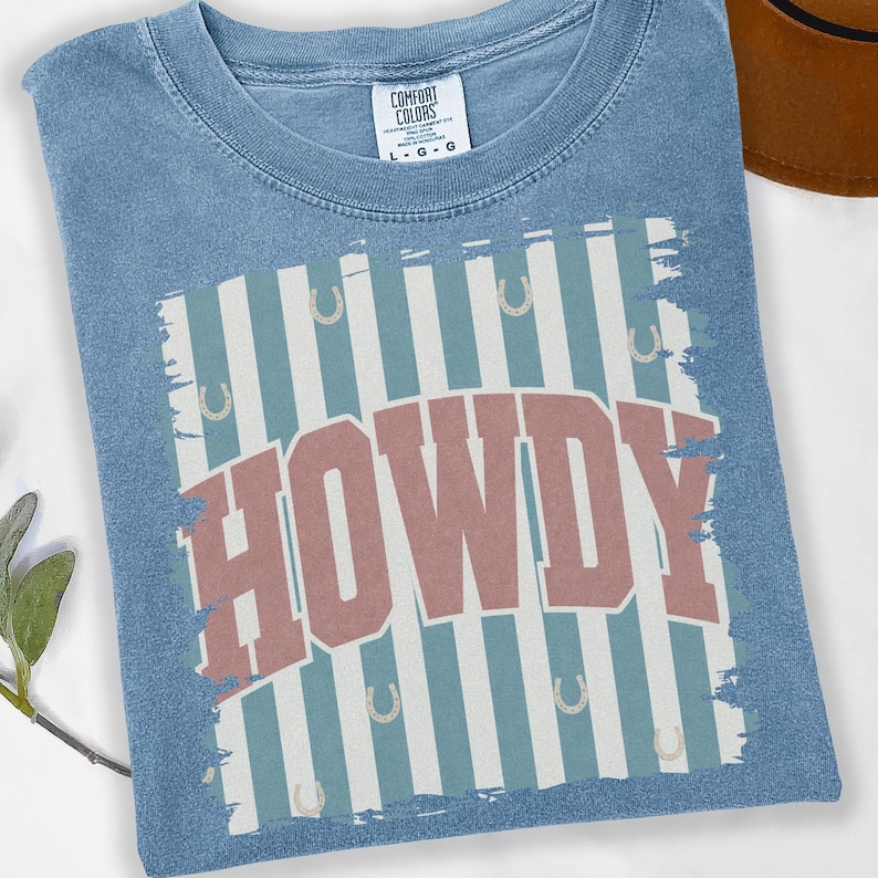 Peut inclure: T-shirt bleu clair avec le mot "HOWDY" en rouge sur un fond de rayures verticales blanches et bleu clair. Le t-shirt a un col rond et une &eacute;tiquette "Comfort Colors".