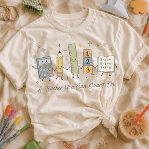 May include: A cream-colored t-shirt featuring a graphic of a smiling calculator, pencil, ruler, numbered blocks, and an open book with math equations. Below the characters, the text reads A Teacher You Can Count On in a cursive script font.