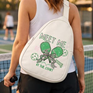 May include: White sling bag with a green and pink pickleball-themed graphic. The graphic includes pickleball paddles, balls, and the text "MEET ME AT THE COURT". The bag has a single strap and a zipper closure.
