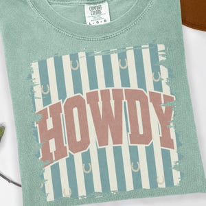 Peut inclure: T-shirt vert clair avec le mot "HOWDY" en rouge et blanc. Le mot est plac&eacute; sur un fond de rayures bleues et blanches avec des fers &agrave; cheval. &Eacute;tiquette Comfort Colors.