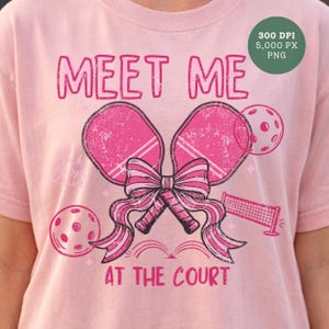 Può includere: T-shirt rosa chiaro con grafica invecchiata. Il design include due racchette da pickleball rosa legate con un fiocco, una pallina da pickleball e una rete. Il testo dice "MEET ME AT THE COURT".