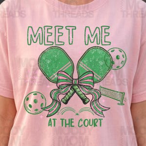 May include: Pink t-shirt with a green and pink pickleball design. The graphic includes two pickleball paddles tied with a bow, two pickleballs, and a net. The text "MEET ME AT THE COURT" is printed in green.
