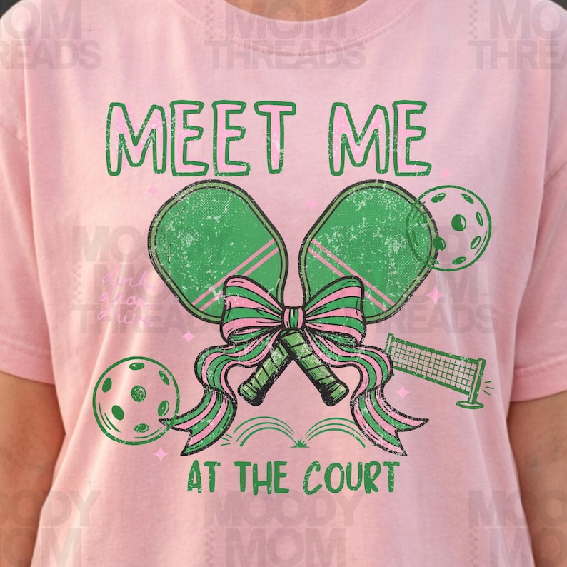 May include: Pink t-shirt with a green and pink pickleball design. The graphic includes two pickleball paddles tied with a bow, two pickleballs, and a net. The text "MEET ME AT THE COURT" is printed in green.