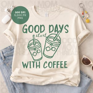 May include: Cream-colored t-shirt with the text "GOOD DAYS start WITH COFFEE" in green. The design includes two green coffee cup illustrations with ice cubes and straws. The shirt is folded on a pair of blue jeans.