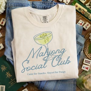 K&ouml;nnte beinhalten: Cremefarbenes T-Shirt mit Cocktail-Grafik und der Aufschrift "Mahjong Social Club". Das Shirt liegt auf einer Jeans, mit Mahjong-Steinen und einem gr&uuml;nen Buch im Hintergrund.