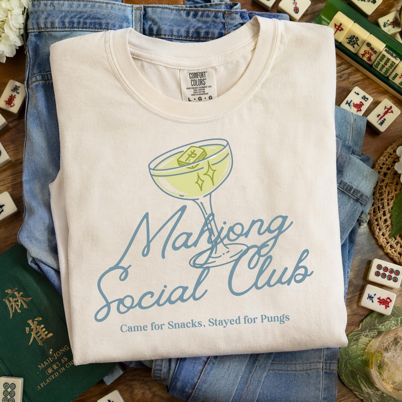 K&ouml;nnte beinhalten: Cremefarbenes T-Shirt mit Cocktail-Grafik und der Aufschrift "Mahjong Social Club". Das Shirt liegt auf einer Jeans, mit Mahjong-Steinen und einem gr&uuml;nen Buch im Hintergrund.