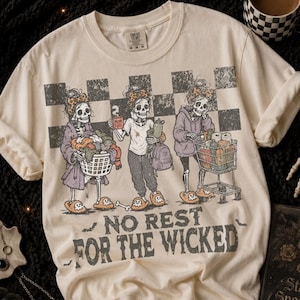 May include: A cream-colored t-shirt featuring a graphic of three skeletons in leopard-print headbands. One holds a laundry basket, one a mug labeled MOM FUEL, and one a shopping cart. The text NO REST FOR THE WICKED is printed below in black.