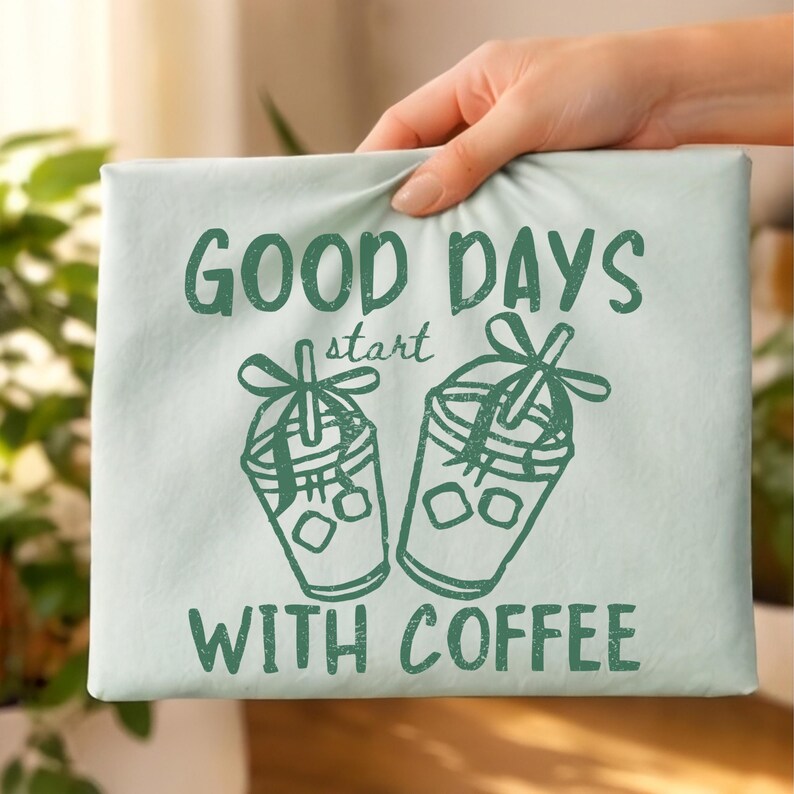 K&ouml;nnte beinhalten: Hellblaue Leinwand mit gr&uuml;nem Text "GOOD DAYS start WITH COFFEE". Zwei gr&uuml;ne Illustrationen von Eiskaffee befinden sich unter dem Text. Die Leinwand wird von einer Hand gehalten.