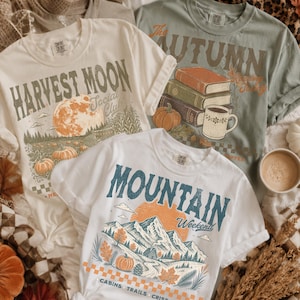 May include: Three short-sleeve t-shirts featuring graphic prints. One cream shirt displays a moon and pumpkins with the text Harvest Moon Social Club. One sage green shirt shows books, a pumpkin, and a mug with the text The Autumn Reading Today. One white shirt depicts mountains and trees with the text Mountain Weekend.