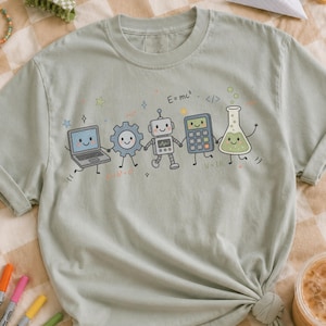 Può includere: Una t-shirt verde salvia con una stampa grafica di cinque personaggi scientifici antropomorfi sorridenti: un portatile, un ingranaggio, un robot, una calcolatrice e un becher. Il design include piccole stelle e formule matematiche come E=mc².